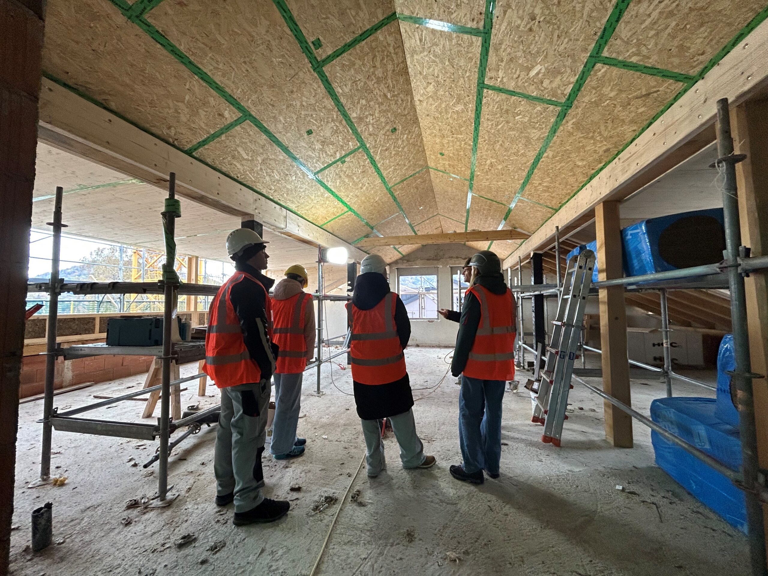 Eine Gruppe von vier Personen mit Schutzhelmen und orangefarbenen Westen steht in einem grossen Gebäude im Bau. Der Raum hat freiliegende Holzbalken und Gerüste. Licht fällt durch grosse Fenster am anderen Ende des Raums ein.