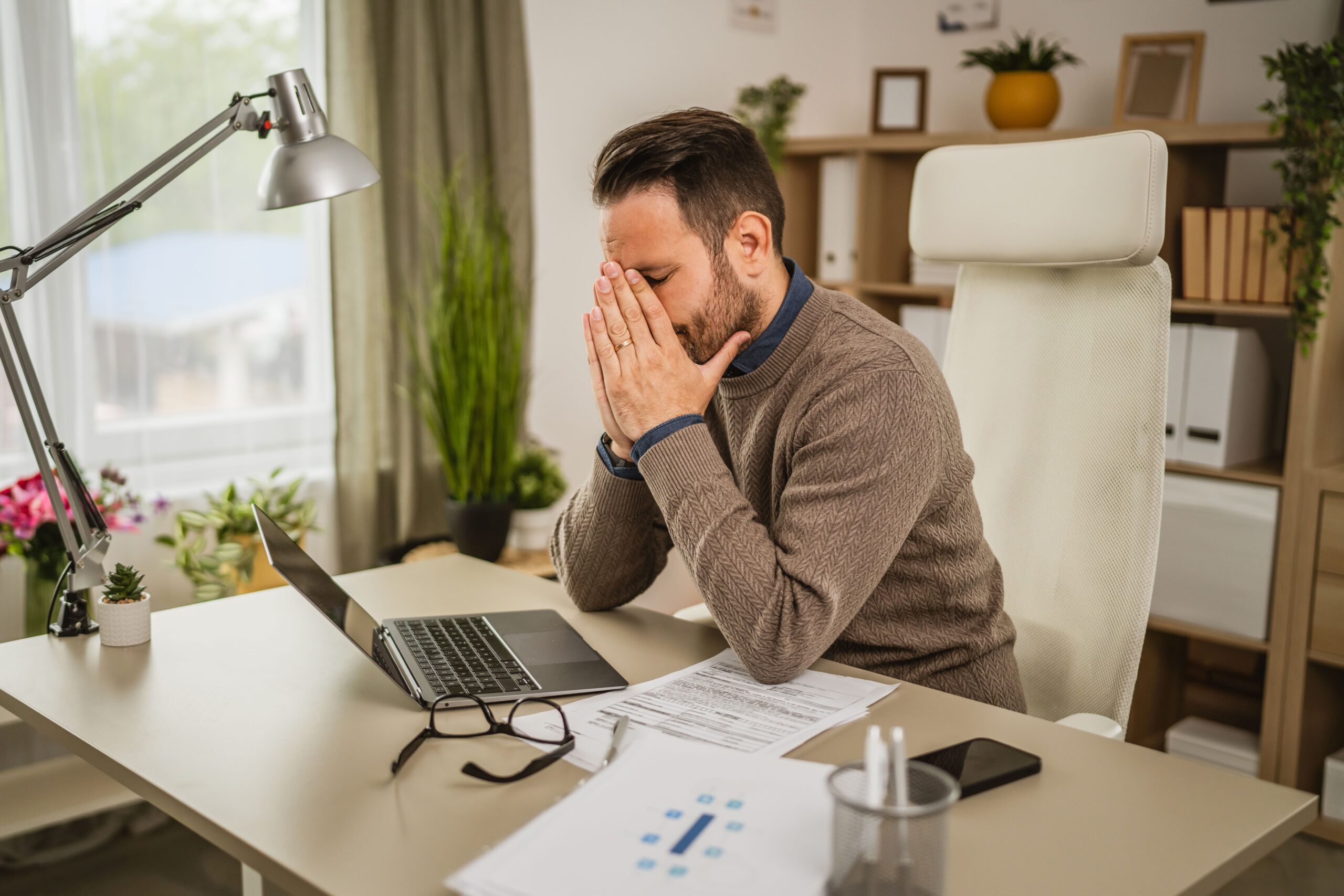 Erschöpfter Mann sitzt am Bürotisch vor dem Laptop und reibt sich die Augen.